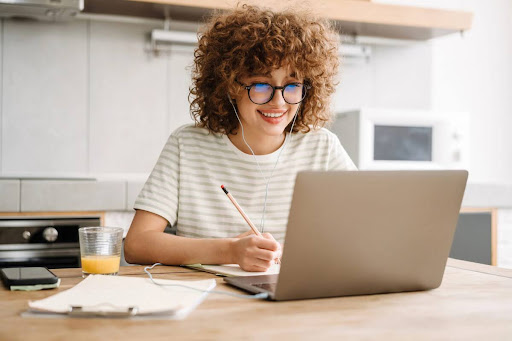 A young woman uses a laptop and takes notes with earbuds in